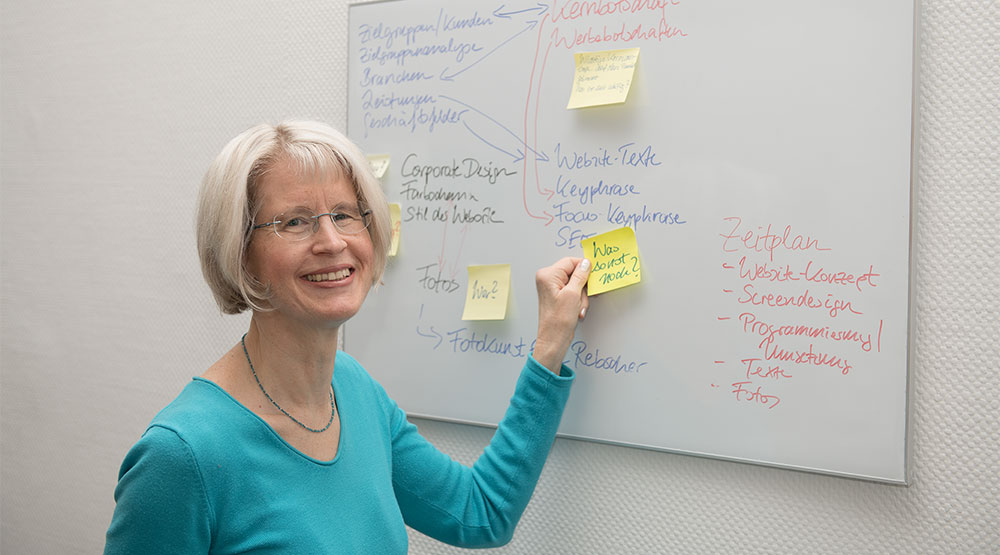 Frau mit blonden Haaren in türkisfarbenem Pullover klebt Notizzettel an Whiteboard, an dem schon weitere Notizzettel mit handschriftlichen Notizen hängen